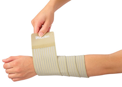 A person's arm and hand are shown wearing and adjusting a beige elastic compression sleeve with multiple pleated bands. The brand name 'Mueller' is printed on the top cuff. - okotoks hc pharmacy