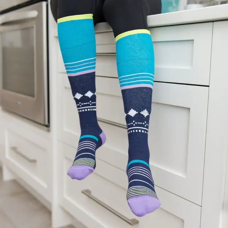 A person sitting on white kitchen cabinets wearing knee-high patterned socks. The socks have bright turquoise and navy sections with white geometric motifs and purple toes and heels. - okotoks hc pharmacy