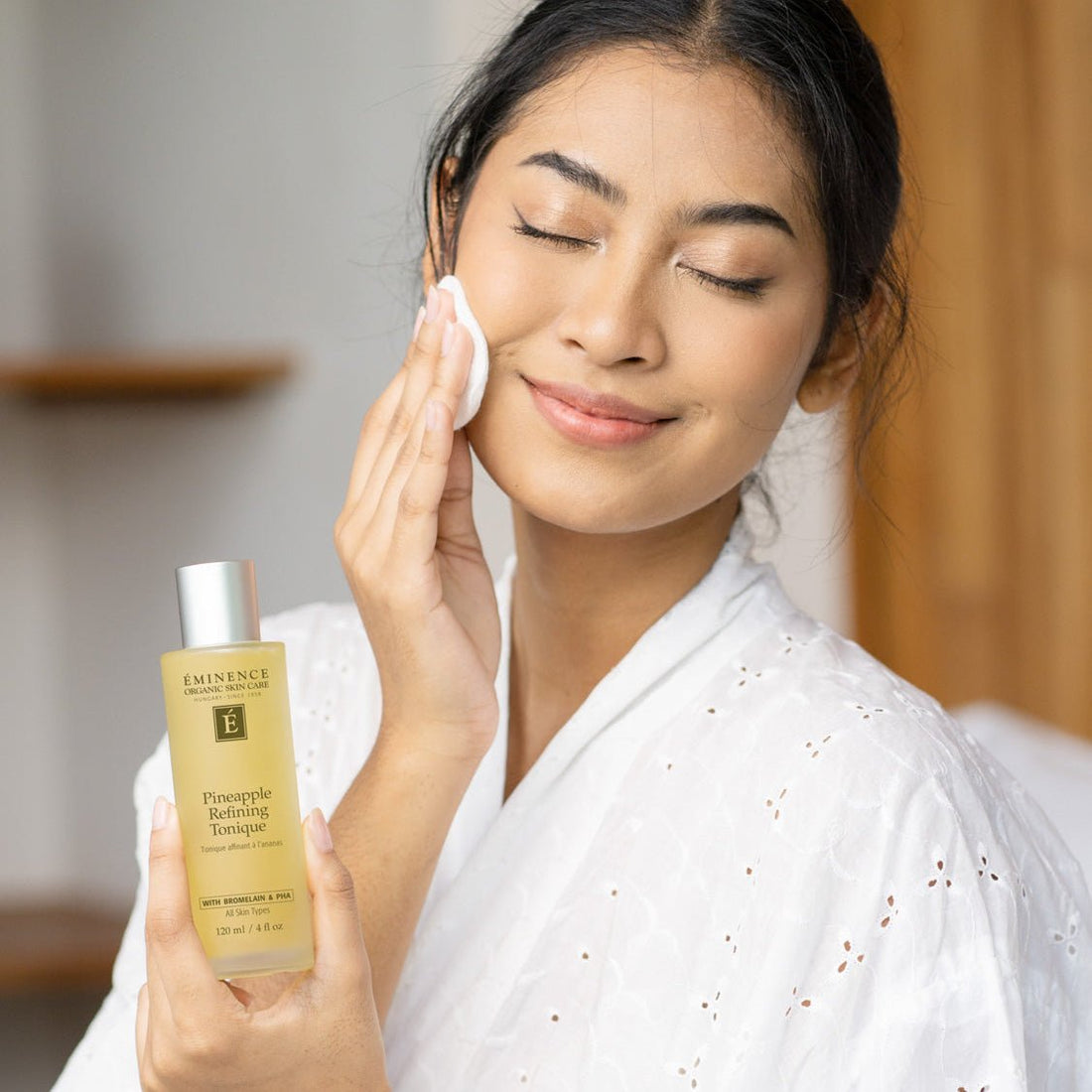 Woman applying facial cleanser with a bottle of Eminence Pineapple Refining Tonique in a bathroom setting. - okotoks hc pharmacy