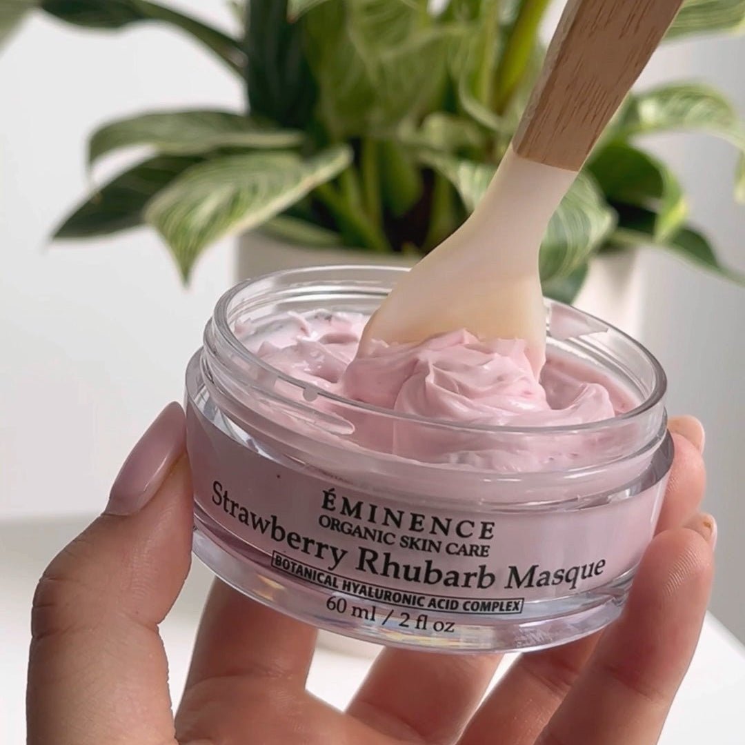 Hand holding a jar of Eminence Strawberry Khubarb Masque with a wooden applicator, plant in the background. - okotoks hc pharmacy