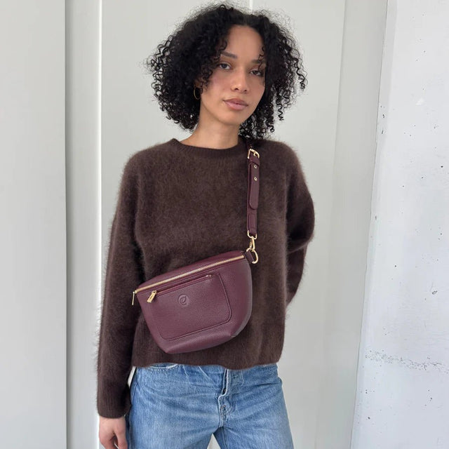Woman wearing a brown sweater and blue jeans with a wine crossbody bag against a white wall. - okotoks hc pharmacy