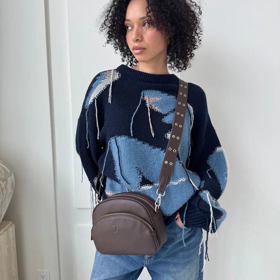 Woman wearing a blue and black patterned sweater with a brown bag, standing against a white wall. - okotoks hc pharmacy