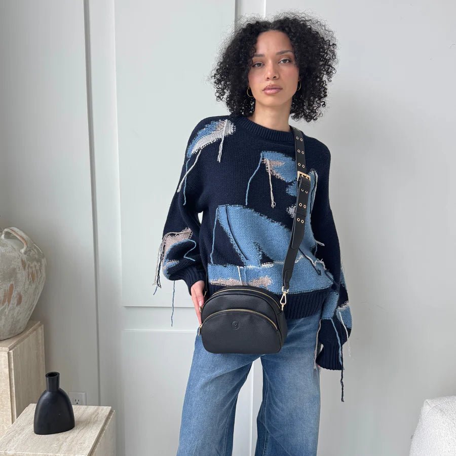 Woman wearing a dark blue sweater with abstract patterns and light blue jeans, holding a black handbag. - okotoks hc pharmacy