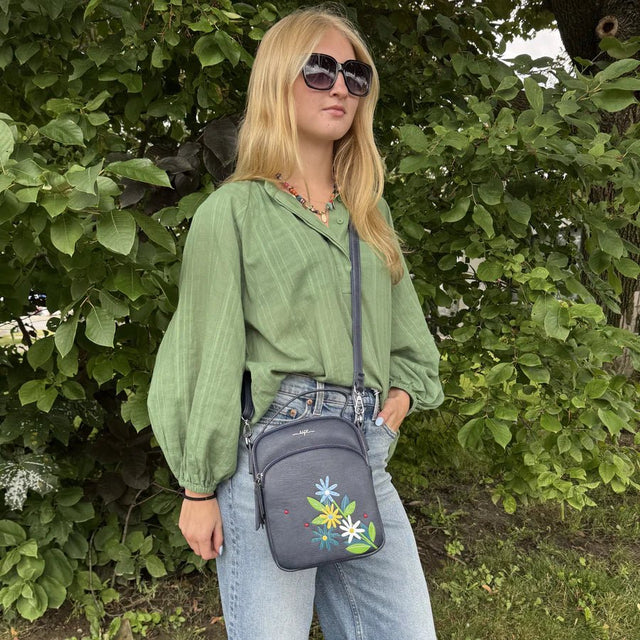 Woman wearing a green shirt and sunglasses, holding a gray crossbody bag with floral designs, standing in front of green foliage. - okotoks hc pharmacy