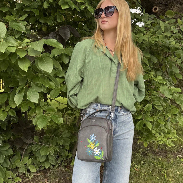 Woman wearing a green shirt and sunglasses, holding a gray crossbody bag with floral design, standing in front of green foliage. - okotoks hc pharmacy