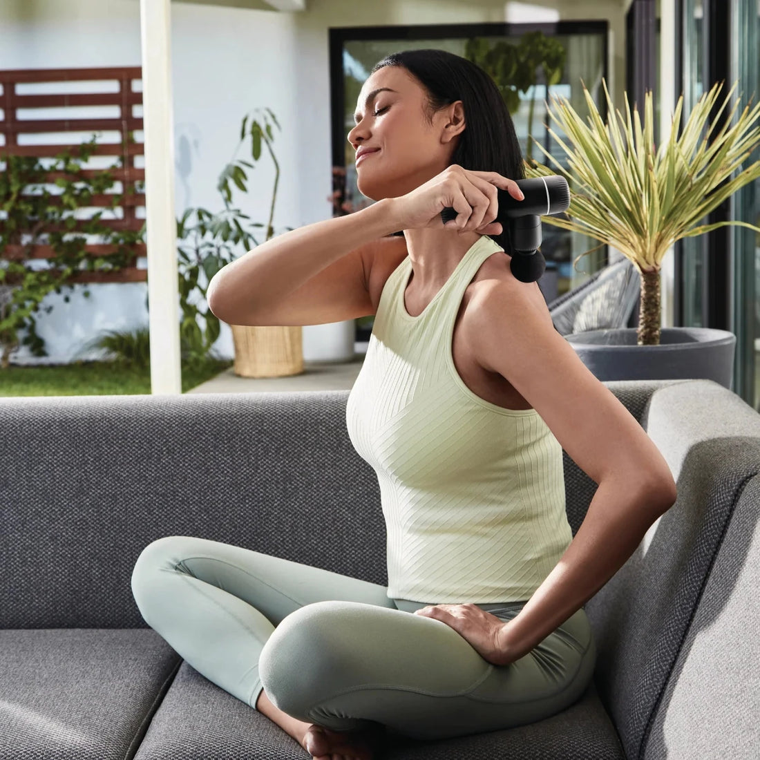 Woman using a handheld massager on her arm while sitting on a couch outdoors. - okotoks hc pharmacy