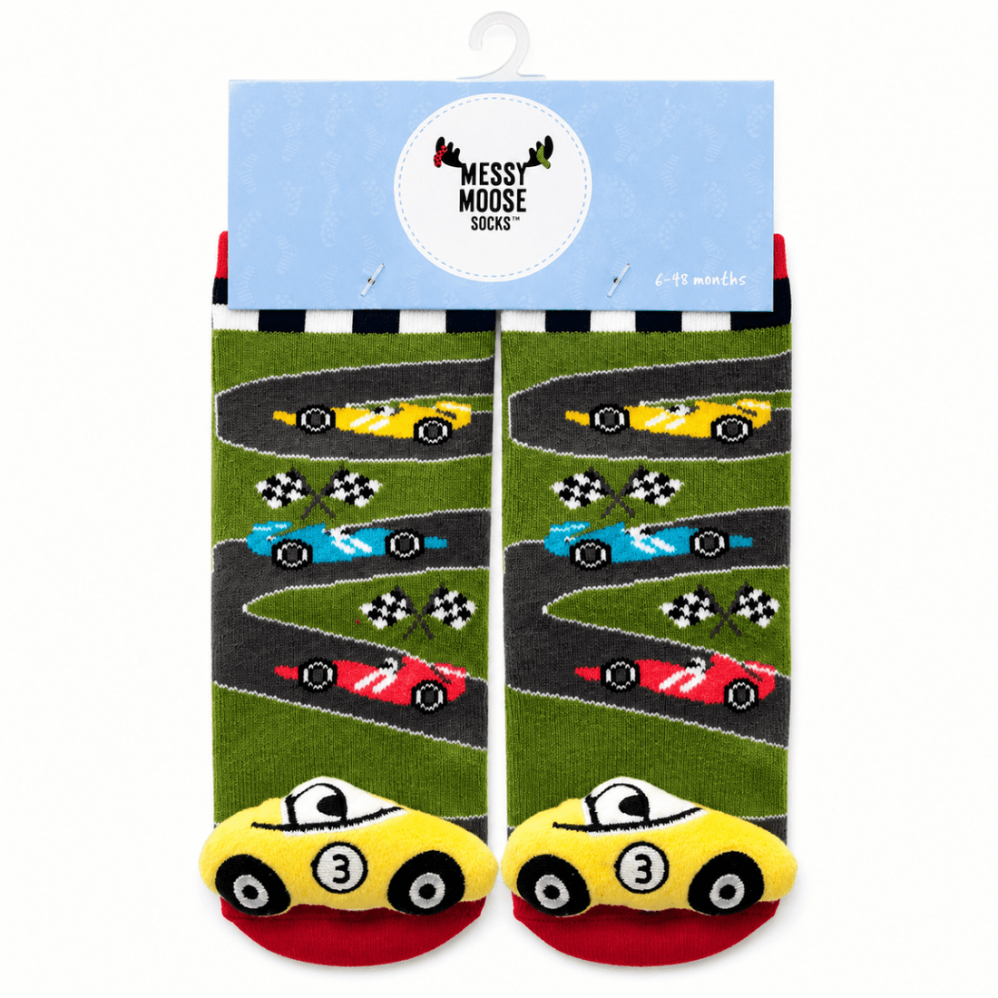 A pair of children's socks with a racing theme, hanging from a light blue cardboard hanger. The socks feature a green grass-like background with winding gray tracks, colorful race cars (yellow, blue, red), and checkered flags, with yellow cars at the toes on a red footbed. - okotoks hc pharmacy
