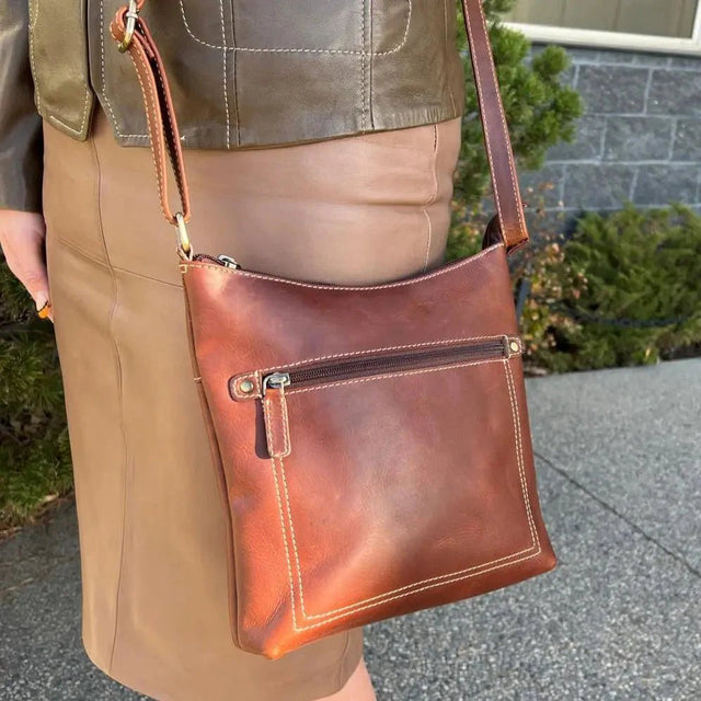 Brown leather crossbody bag held by a person wearing a beige skirt. - okotoks hc pharmacy