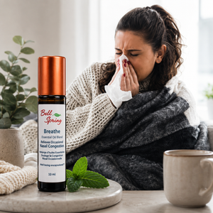 A woman wrapped in a gray blanket and wearing a knit sweater sits indoors, holding a tissue to her nose. In the foreground, a small essential oil bottle labeled Bell Spring Breathe sits on a tray beside mint leaves and a mug. - okotoks hc pharmacy