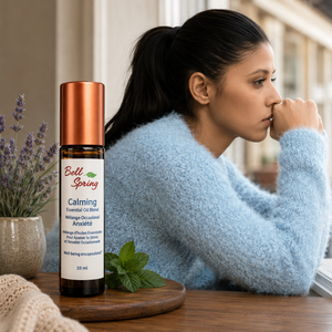A woman in a fuzzy light blue sweater sits at a wooden table, looking out a window. In the foreground, a bottle of Bell Spring Calming Essential Oil Blend with a copper-colored cap stands on a small wooden tray next to mint leaves and lavender. - okotoks hc pharmacy