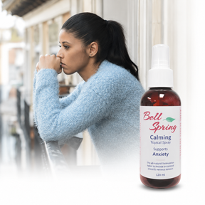A woman in a light blue fuzzy sweater leans against a white wall, looking pensive, with a bottle of Bell Spring Calming Tropical Spray prominently shown in the foreground on a white surface. - okotoks hc pharmacy