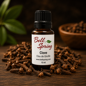 A 15 ml bottle of Bell Spring Clove essential oil, surrounded by loose whole cloves on a dark wooden surface with a blurred background showing green leaves and a bowl of cloves. - okotoks hc pharmacy
