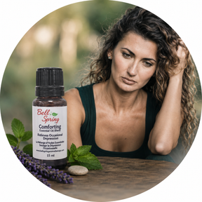 A woman with curly brown hair sits at a wooden table with a bottle of Bell's Spring Comforting Essential Oil Blend, surrounded by green leaves, purple lavender, and a stone, in a circular frame with a blurred green background. - okotoks hc pharmacy
