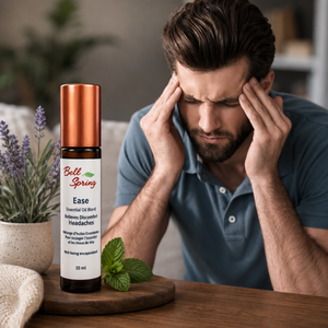 A 20 ml bottle of Bell Spring Ease Essential Oil Blend sits on a wooden tray on a table, next to mint leaves and lavender. In the background, a man in a blue polo shirt holds his head in his hands in apparent discomfort, representing a headache or stress scenario. - okotoks hc pharmacy