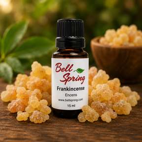 A small amber glass bottle of Bell Spring Frankincense with a black cap and white label, surrounded by loose yellow-orange frankincense buds on a wooden surface with a blurred green background. - okotoks hc pharmacy