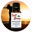 A 15 ml amber glass essential oil bottle with a black cap and a white label reading 'Bell Spring' and 'Raise Your Spirit', centered over a circular beach sunset background with a silhouetted person. - okotoks hc pharmacy
