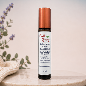 A 10 ml roll-on bottle of Belle Spring 'Raise Your Spirit' essential oil blend, standing on a round marble tray next to a folded beige cloth and lavender sprigs against a soft blue and beige gradient background. - okotoks hc pharmacy