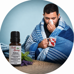 A man wrapped in a blue striped blanket and holding a mug appears in a circular lifestyle scene next to a small Bell Spring Relieve essential oil bottle with lavender sprigs, on a light blue background. - okotoks hc pharmacy