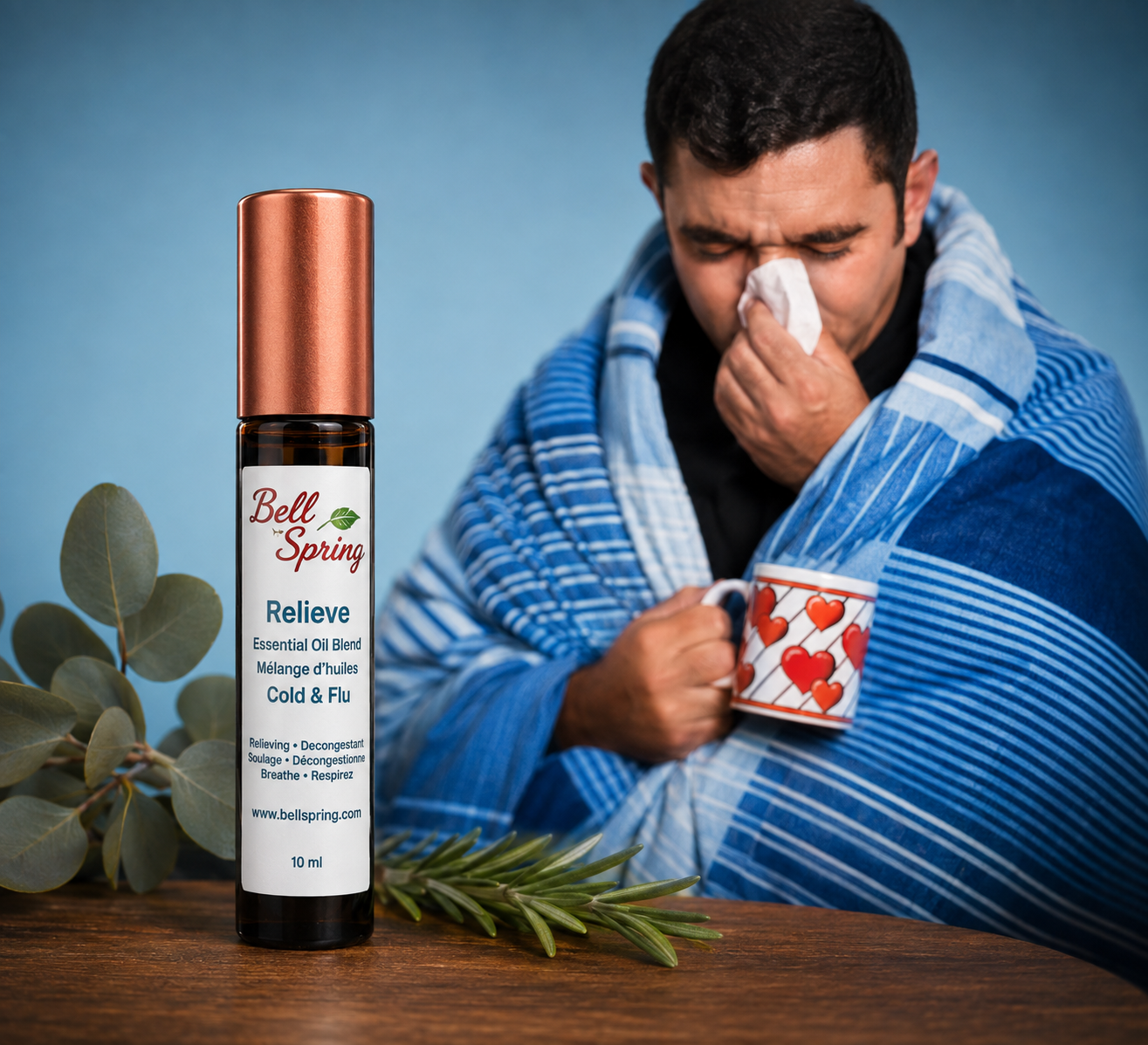A bottle of Bell Spring Relieve essential oil blend sits on a wooden table in the foreground, while a man wrapped in a blue striped blanket and holding a mug with a heart pattern sneezes in the background against a blue backdrop. - okotoks hc pharmacy