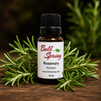 A 15 ml bottle of Bell Spring Rosemary essential oil, displayed on a rustic wooden surface and surrounded by fresh green rosemary sprigs. The bottle has a black cap and a white label with red and green text. - okotoks hc pharmacy