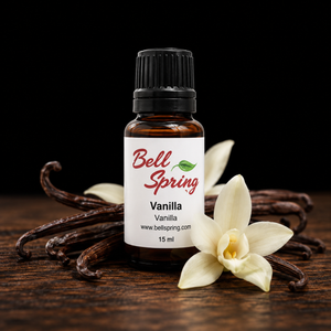 A 15 ml amber glass bottle of Bell Spring Vanilla essential oil with a white label and black cap, styled on a dark wooden surface with vanilla beans and white vanilla flowers against a black background. - okotoks hc pharmacy