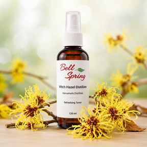 Bell Spring Witch Hazel Distillate Refreshing Toner in a 120 ml amber glass spray bottle, styled with yellow witch hazel flowers on a wooden surface against a soft, blurred green bokeh background. - okotoks hc pharmacy