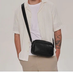 A man wearing a white short-sleeve button-up shirt and beige pants, carrying a black leather crossbody bag across his chest. He is standing against a plain light gray studio background. - okotoks hc pharmacy