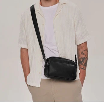 A man wearing a white short-sleeve button-up shirt and beige pants, carrying a black leather crossbody bag across his chest. He is standing against a plain light gray studio background. - okotoks hc pharmacy