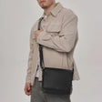 A man wearing a beige overshirt and gray jeans carries a black leather crossbody messenger bag against his side. - okotoks hc pharmacy