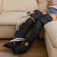 A person reclining on a beige sofa wearing a black electric leg massage device with white cords and a small control unit resting on the couch. - okotoks hc pharmacy
