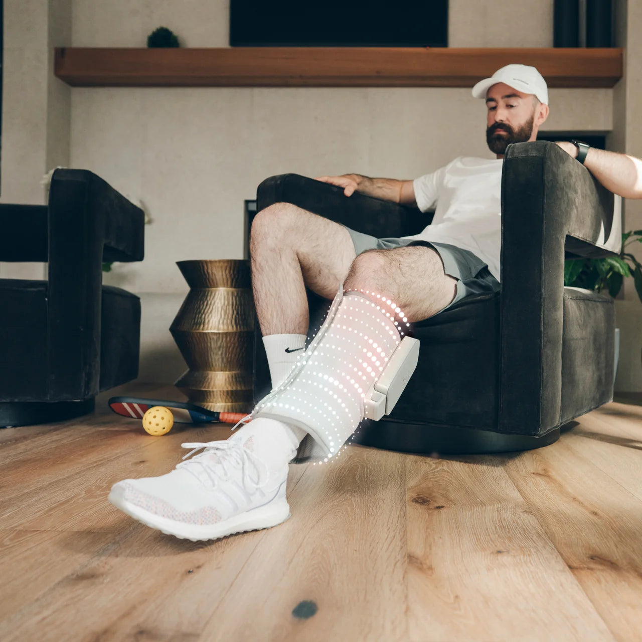A man sits in a dark armchair in a living room while wearing a white and gray electronic leg stimulator device on his lower leg, with a white Nike Air Max sneaker visible. - okotoks hc pharmacy
