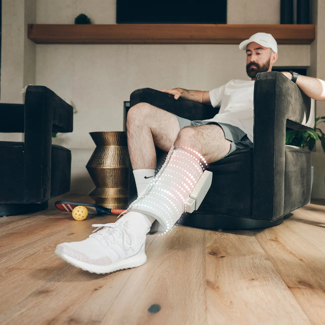 A man sits in a dark armchair in a living room while wearing a white and gray electronic leg stimulator device on his lower leg, with a white Nike Air Max sneaker visible. - okotoks hc pharmacy