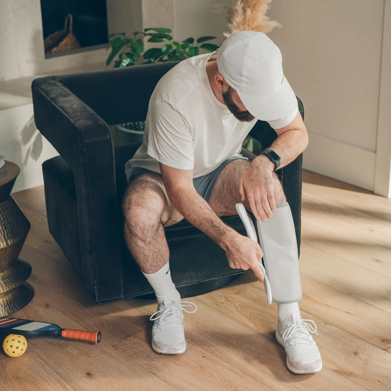 A man in a white t-shirt, shorts, and white sneakers sits on a black armchair in a bright room, applying a white compression bandage to his lower leg. - okotoks hc pharmacy
