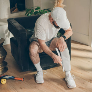A man in a white t-shirt, shorts, and white sneakers sits on a black armchair in a bright room, applying a white compression bandage to his lower leg. - okotoks hc pharmacy