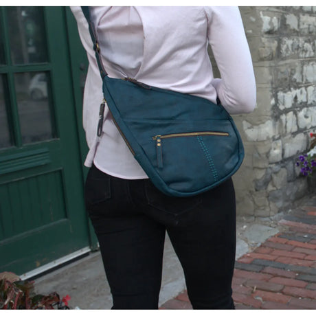 A woman wearing a light pink long-sleeve top and black pants carries a teal blue crossbody bag with gold-toned hardware in an outdoor setting. - okotoks hc pharmacy