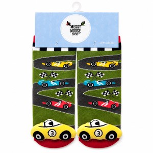 A pair of children's socks with a racing theme, hanging from a light blue cardboard hanger. The socks feature a green grass-like background with winding gray tracks, colorful race cars (yellow, blue, red), and checkered flags, with yellow cars at the toes on a red footbed. - okotoks hc pharmacy