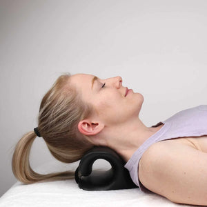 A woman lying down with her head resting on a black curved neck pillow, wearing a light purple tank top against a plain light gray background. - okotoks hc pharmacy