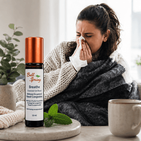A woman wrapped in a gray blanket and wearing a knit sweater sits indoors, holding a tissue to her nose. In the foreground, a small essential oil bottle labeled Bell Spring Breathe sits on a tray beside mint leaves and a mug. - okotoks hc pharmacy