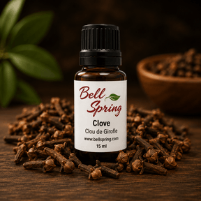 A 15 ml bottle of Bell Spring Clove essential oil, surrounded by loose whole cloves on a dark wooden surface with a blurred background showing green leaves and a bowl of cloves. - okotoks hc pharmacy