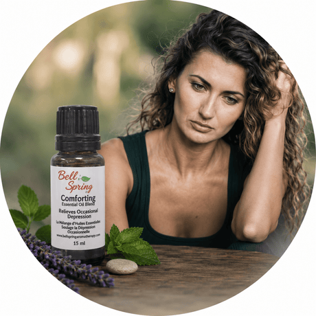 A woman with curly brown hair sits at a wooden table with a bottle of Bell's Spring Comforting Essential Oil Blend, surrounded by green leaves, purple lavender, and a stone, in a circular frame with a blurred green background. - okotoks hc pharmacy