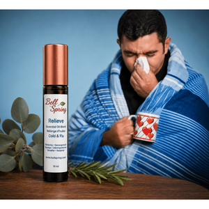 A bottle of Bell Spring Relieve essential oil blend sits on a wooden table in the foreground, while a man wrapped in a blue striped blanket and holding a mug with a heart pattern sneezes in the background against a blue backdrop. - okotoks hc pharmacy