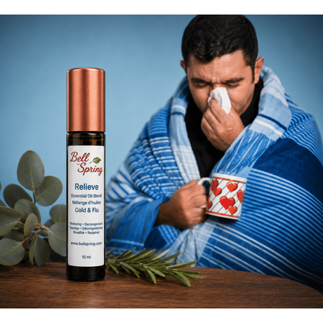 A bottle of Bell Spring Relieve essential oil blend sits on a wooden table in the foreground, while a man wrapped in a blue striped blanket and holding a mug with a heart pattern sneezes in the background against a blue backdrop. - okotoks hc pharmacy