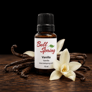 A 15 ml amber glass bottle of Bell Spring Vanilla essential oil with a white label and black cap, styled on a dark wooden surface with vanilla beans and white vanilla flowers against a black background. - okotoks hc pharmacy