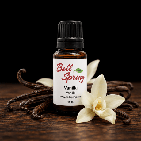 A 15 ml amber glass bottle of Bell Spring Vanilla essential oil with a white label and black cap, styled on a dark wooden surface with vanilla beans and white vanilla flowers against a black background. - okotoks hc pharmacy