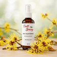 Bell Spring Witch Hazel Distillate Refreshing Toner in a 120 ml amber glass spray bottle, styled with yellow witch hazel flowers on a wooden surface against a soft, blurred green bokeh background. - okotoks hc pharmacy