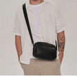A man wearing a white short-sleeve button-up shirt and beige pants, carrying a black leather crossbody bag across his chest. He is standing against a plain light gray studio background. - okotoks hc pharmacy