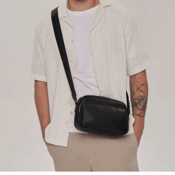 A man wearing a white short-sleeve button-up shirt and beige pants, carrying a black leather crossbody bag across his chest. He is standing against a plain light gray studio background. - okotoks hc pharmacy