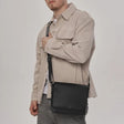 A man wearing a beige overshirt and gray jeans carries a black leather crossbody messenger bag against his side. - okotoks hc pharmacy