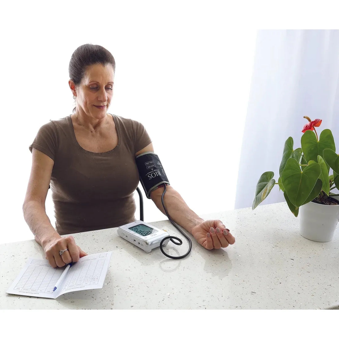 Woman taking her blood pressure at home with a device and recording it in a notebook. - okotoks hc pharmacy