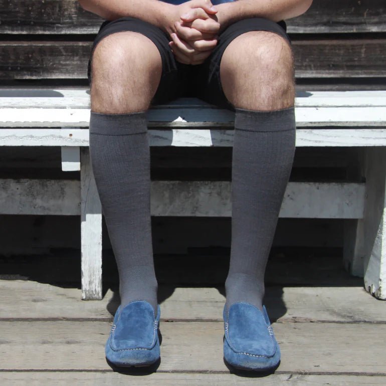 A person seated on a weathered wooden bench wearing dark gray knee-high socks and blue slip-on shoes, photographed outdoors on a wooden deck. - okotoks hc pharmacy